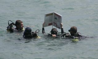 Journalists divers exhibit a newspaper, laid out under water