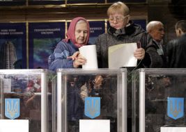 Пожилой женщине помогают бросить бюллетень для голосования в урну