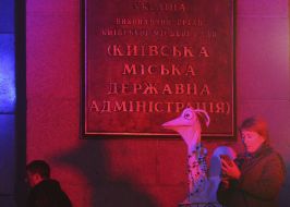An actress with a bird costume on the background of a plaque bearing the inscription KGGA