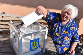 Пожилая женщина бросает бюллетень в переносную урну для голосования