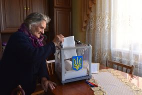 Пожилая женщина бросает бюллетень в переносную урну для голосования
