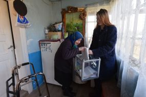 Пожилая женщина бросает бюллетень в переносную урну для голосования