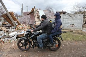 Чоловіки на мотоциклі їдуть повз зруйнований будинок