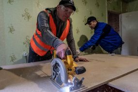 Volunteers cut wood boards to repair broken windows in a house damaged by a MLRS salvo