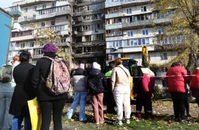 People stand near a fence near a dilapidated and burned residential building after a nighttime drone attack in the Desnyanskyi district of Kyiv
